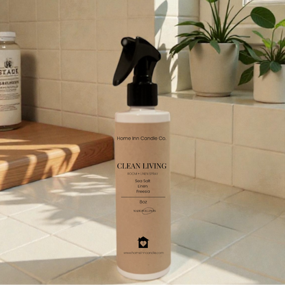 Bottle of 'Clean Living' room spray by Home Inn Candle on a kitchen counter with plants and a window in the background.