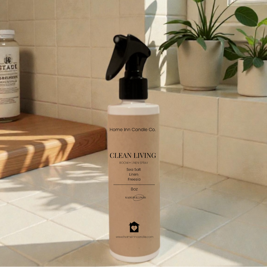 Bottle of 'Clean Living' room spray by Home Inn Candle on a kitchen counter with plants and a window in the background.