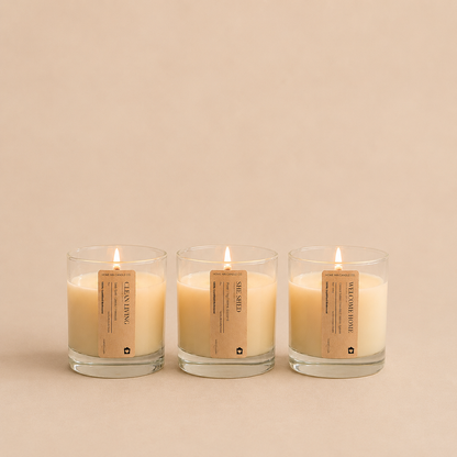 Three lit glass jar candles with beige labels are arranged side by side on a warm beige background. The labels show the brand name 'Home Inn Candle' and scent names including 'She Shed', 'Clean Living', and 'Welcome Home'.