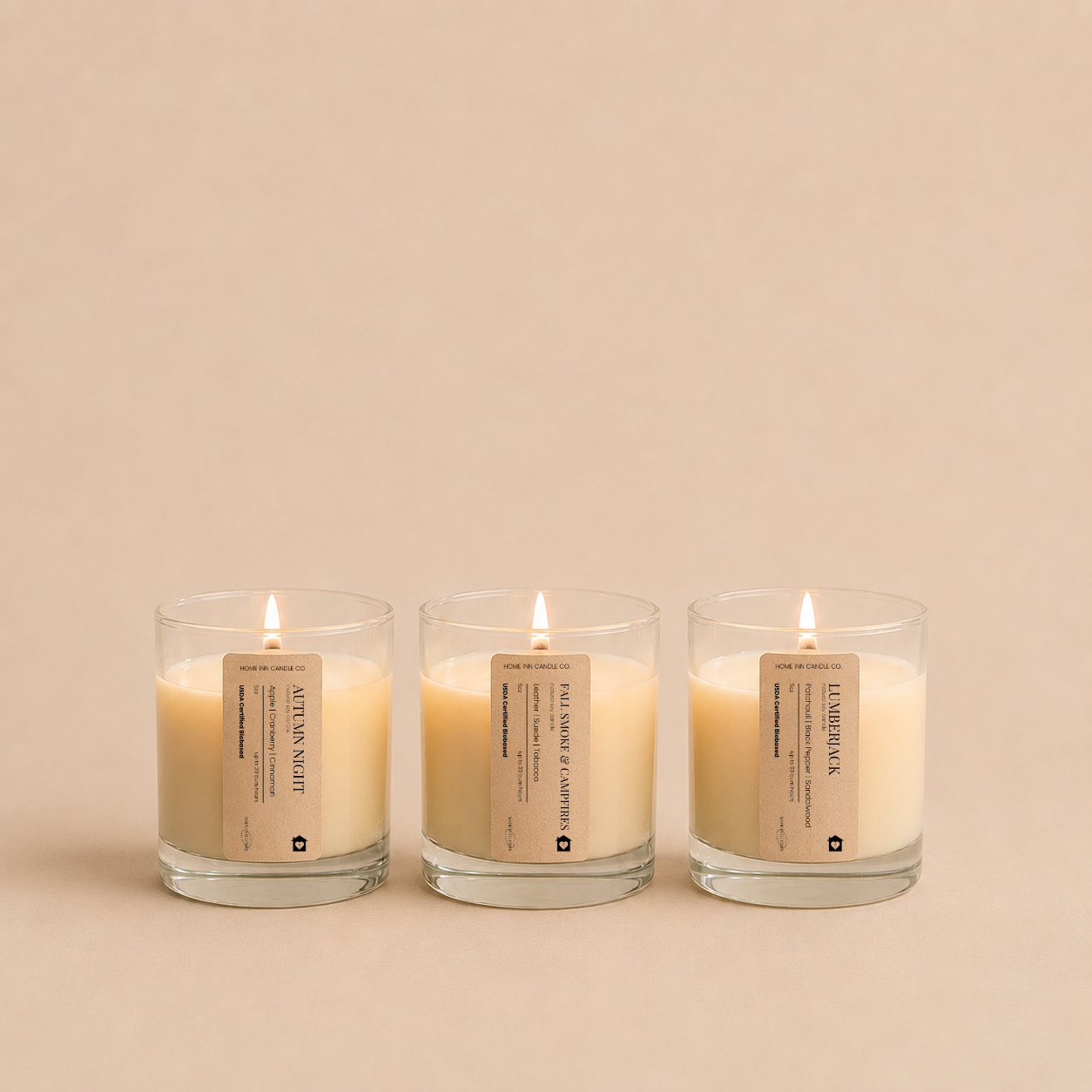 Three lit candles in clear glass jars with beige labels, arranged side by side on a warm beige background. The labels read 'Autumn Night', 'Lumberjack'  and 'Fall Smoke and Campfires' from the Fall Candle Discovery Set by Home Inn Candle