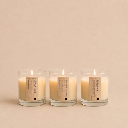 Three lit candles in clear glass jars with beige labels, arranged side by side on a warm beige background. The labels read 'Autumn Night', 'Lumberjack'  and 'Fall Smoke and Campfires' from the Fall Candle Discovery Set by Home Inn Candle