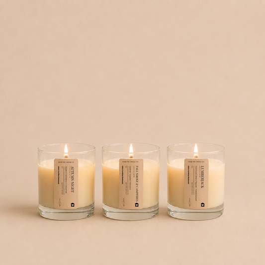 Three lit candles in clear glass jars with beige labels, arranged side by side on a warm beige background. The labels read 'Autumn Night', 'Lumberjack'  and 'Fall Smoke and Campfires' from the Fall Candle Discovery Set by Home Inn Candle