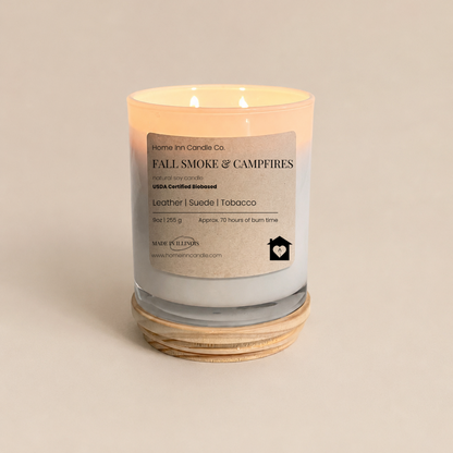 A lit scented candle in a white  glass jar with a cream-colored label reading 'FALL SMOKE & CAMPFIRES' from Home Inn Candle. The candle sits on a  natural wood lid against a solid, warm beige background.
