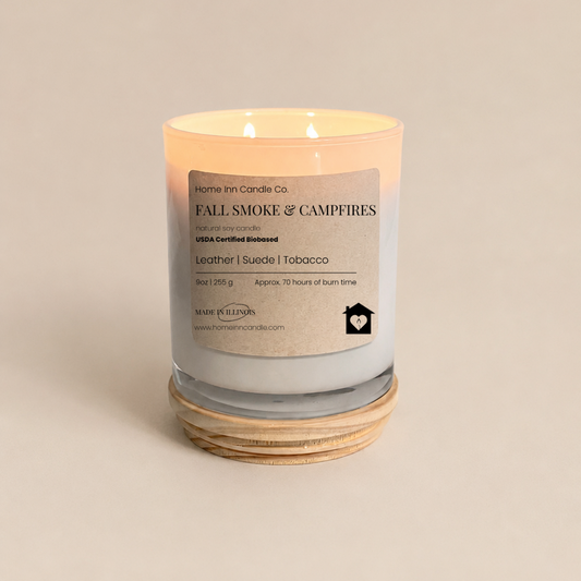 A lit scented candle in a white  glass jar with a cream-colored label reading 'FALL SMOKE & CAMPFIRES' from Home Inn Candle. The candle sits on a  natural wood lid against a solid, warm beige background.