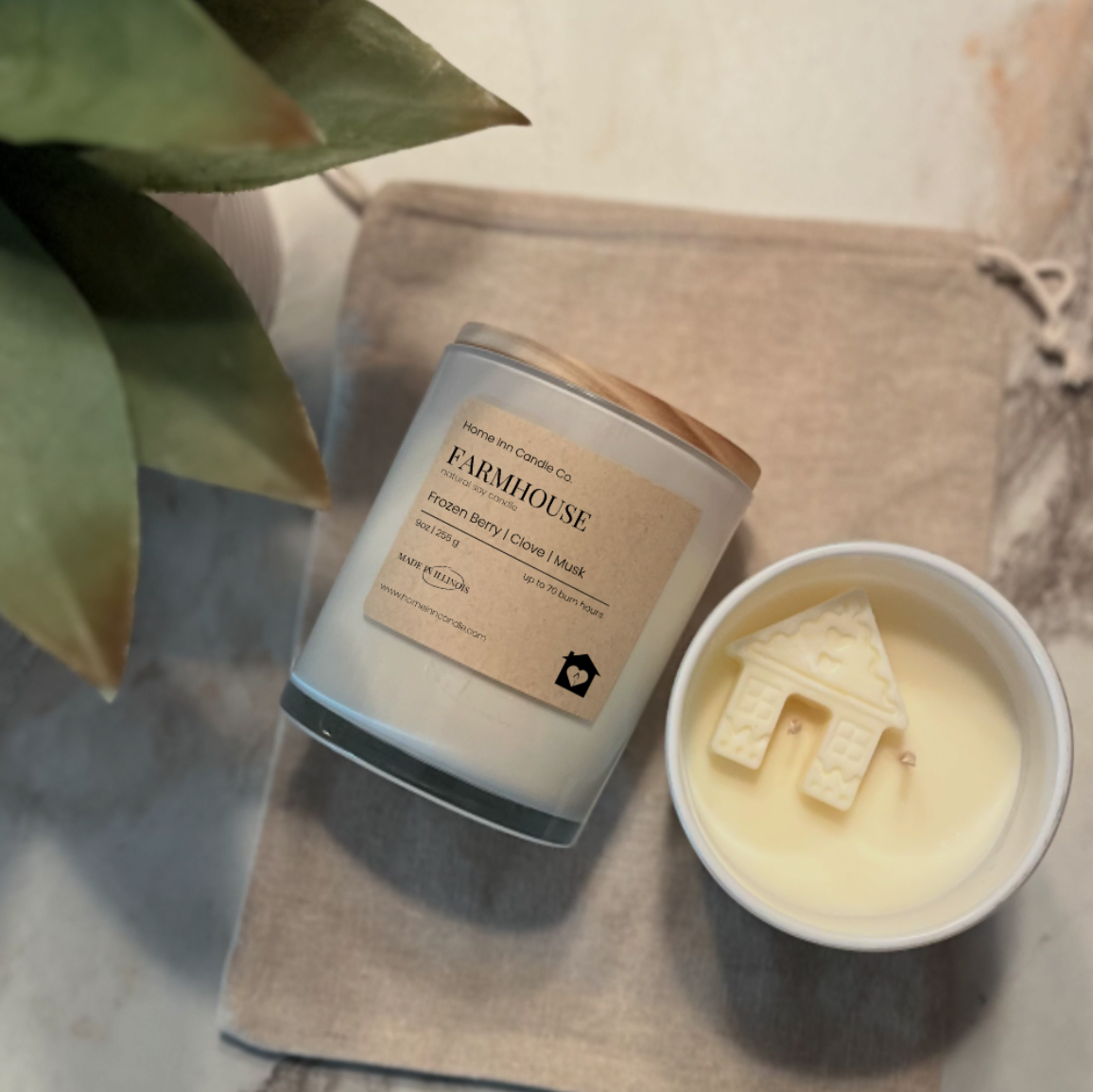 Candle labeled 'Farmhouse' from Home Inn Candle next to a small bowl with a house-shaped candle on a marble surface.