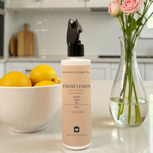 Fresh Lemon room spray from Home Inn Candle co on kitchen counter with fruit bowl of lemons and pink floral arrangement in a vase. 