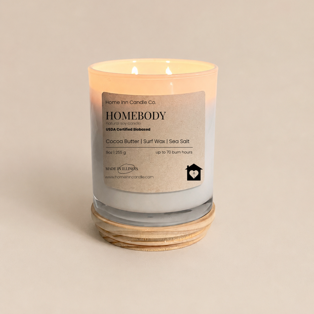 A lit scented candle in a clear glass jar with a cream label reading Homebody by Home Inn Candle Co., sitting on a stack of natural wood lid and beige background.