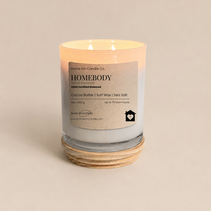 A lit scented candle in a clear glass jar with a cream label reading Homebody by Home Inn Candle Co., sitting on a stack of natural wood lid and beige background.