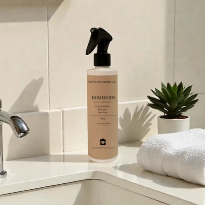 Homebody Room Spray by Home Inn Candle on a bathroom counter with sink and towel in the background