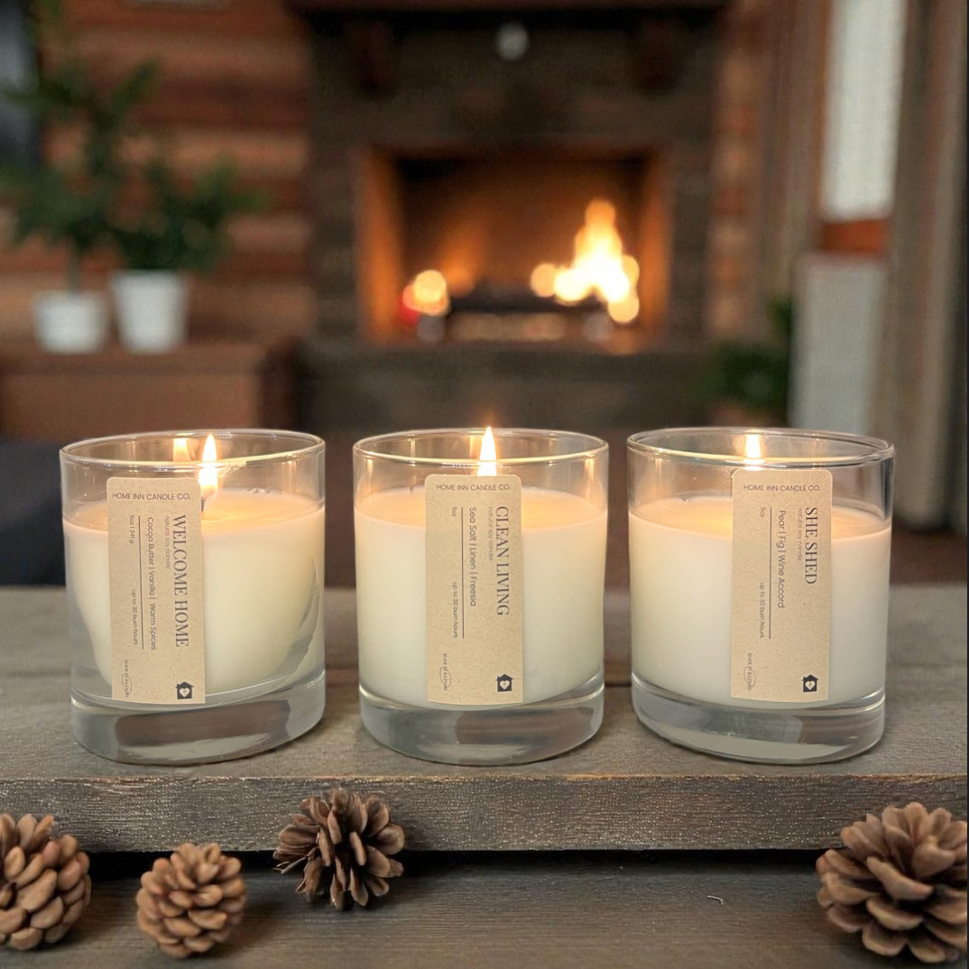 Three lit candles in glass holders on a wooden surface with a fireplace in the background.