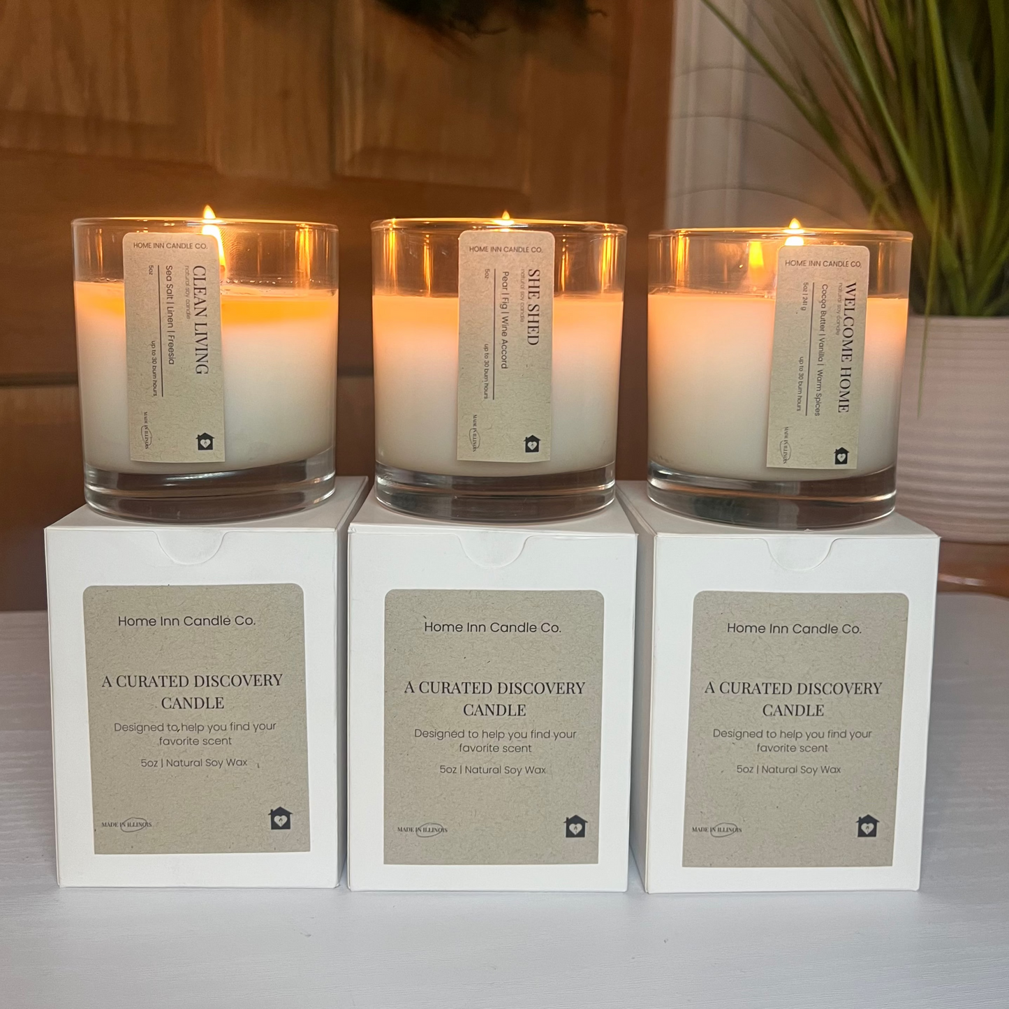 Three lit candles in glass holders on white boxes with labels, set against a neutral background.