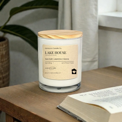 Candle labeled 'Lake House' on a wooden surface with an open book and plant in the background