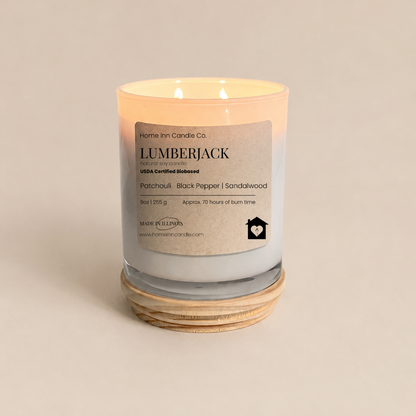 A lit double-wick candle in a WHITE glass jar with a cream label reading 'LUMBERJACK'From Home Inn Candle Co.' The candle sits on a natural wood lid against a warm beige background.