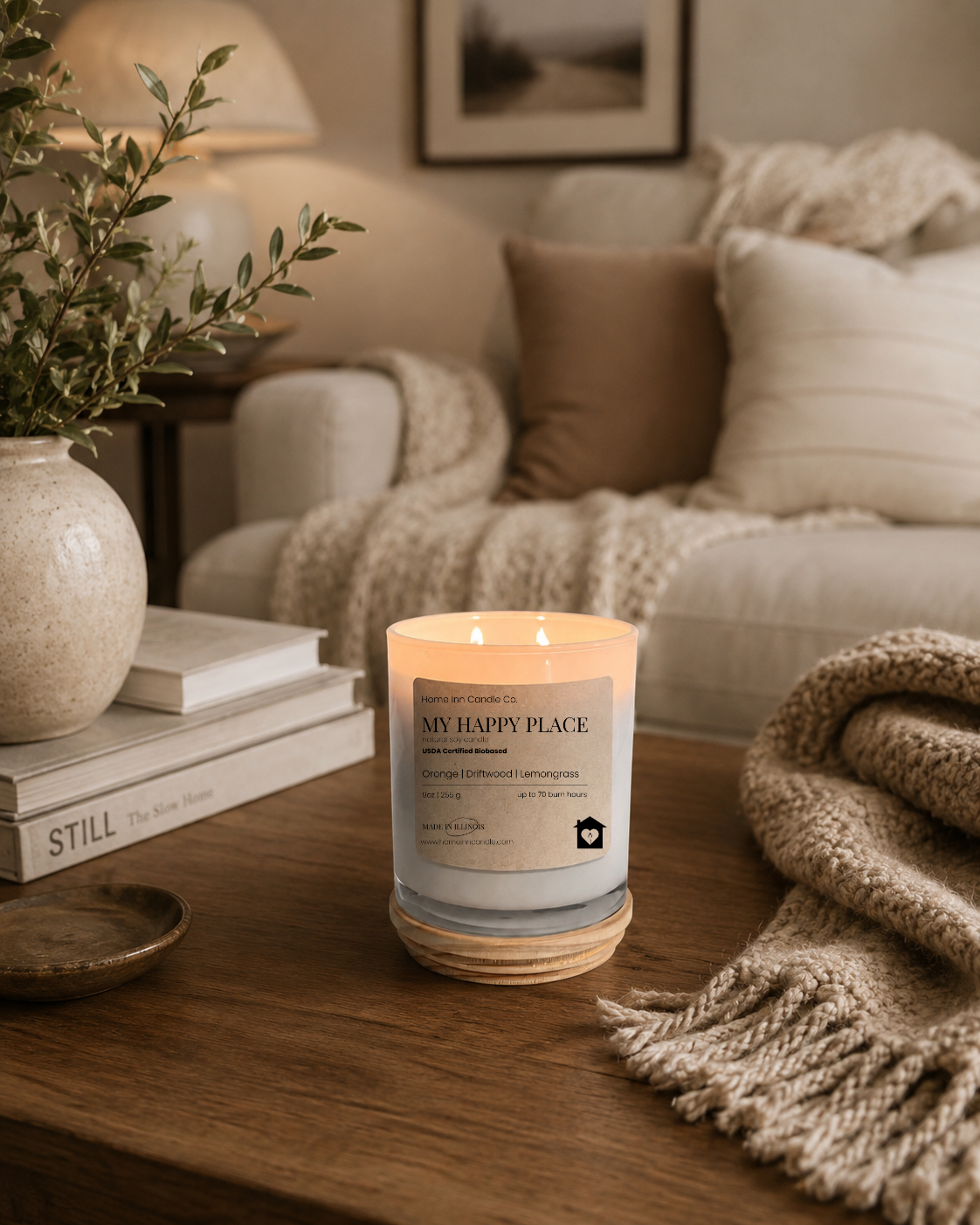 A lit scented candle in a white glass jar with a cream label reading 'My Happy Place' from Home Inn Candle sits on a wooden table in a cozy living room setting with a sofa, throw blanket, and books in the background.