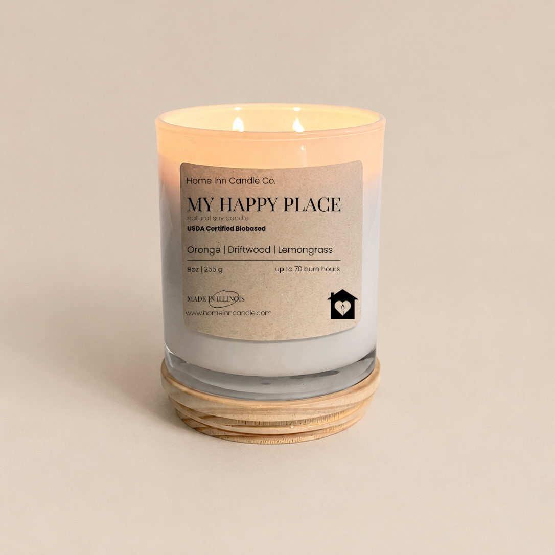 A lit scented candle in a clear glass jar with a cream label reading 'Home Inn Candle Co. MY HAPPY PLACE' and 'Orange | Driftwood | lemongrass,' set on a natural wood lid against a warm beige background.