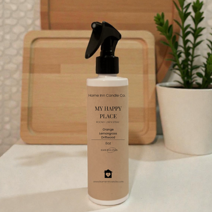 Bottle of 'My Happy Place' spray from Home Inn Candle on a surface with wooden boards and a plant in the background.
