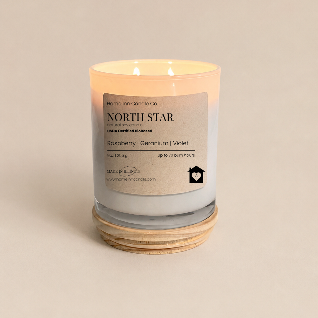 A lit double-wick scented candle in a white glass jar with a white wax fill, resting on a natural wood lid. The label reads 'NORTH STAR' by 'Home Inn Candle Co.