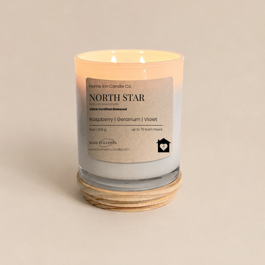 A lit double-wick scented candle in a white glass jar with a white wax fill, resting on a natural wood lid. The label reads 'NORTH STAR' by 'Home Inn Candle Co.