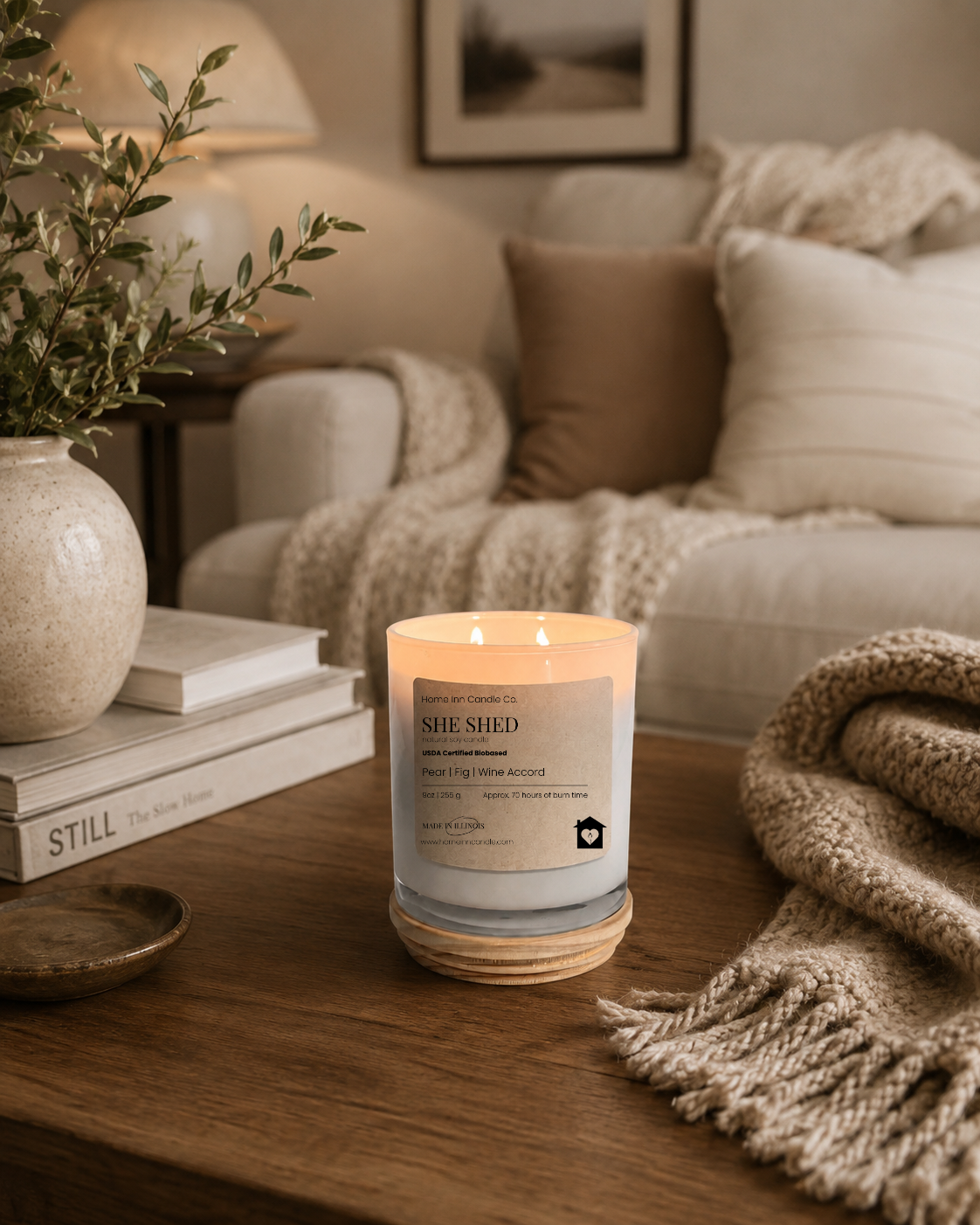 A lit scented candle in a white glass jar with a cream label reading 'She Shed' from Home Inn Candle sits on a wooden table in a cozy living room setting with a sofa, throw blanket, and books in the background.
