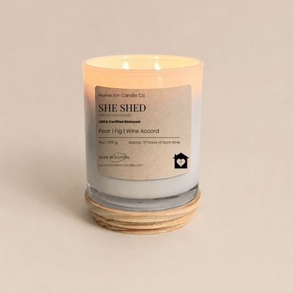 A lit scented candle in a clear glass jar with a white label reading 'SHE SHED' and 'Pear l Fig | Wine Accord', from Home inn Candle