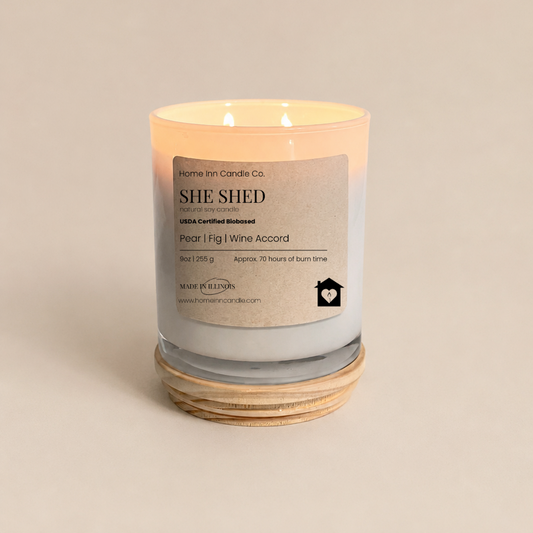 A lit scented candle in a clear glass jar with a white label reading 'SHE SHED' and 'Pear l Fig | Wine Accord', from Home inn Candle