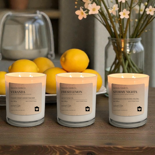 Three candles labeled Fresh Lemon, Veranda, and Stormy Nights from Home Inn Candle Company on a wood table with fruit and floral decor