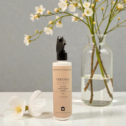 Veranda Room Spray bottle on white surface with floral arrangement in a glass vase