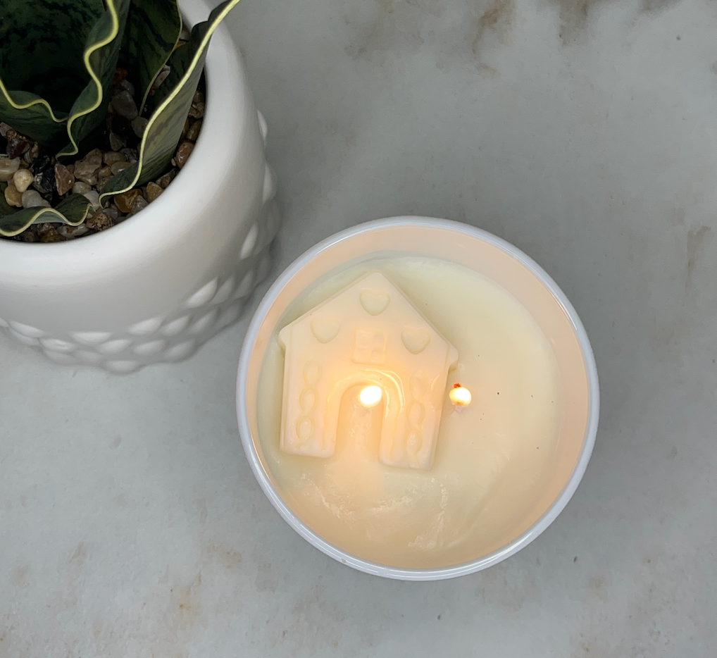 A lit cream-colored scented candle in a white glass jar sits on a light marble surface next to a potted snake plant. The candle features a small house-shaped wax ornament in the center with a warm glow from the two lit wicks. Candle is from Home Inn Candle Company